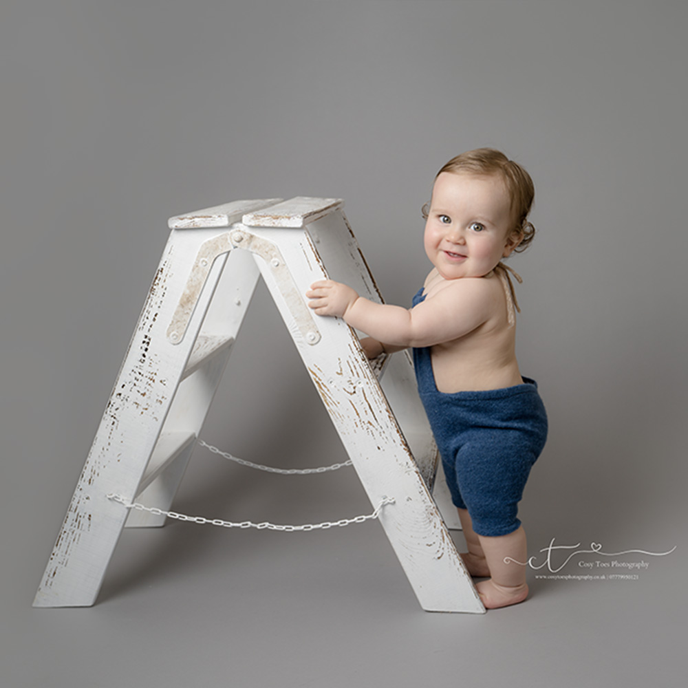 Baby boy standing holding onto a white ladder at his first birthday photoshoot in East Grinstead