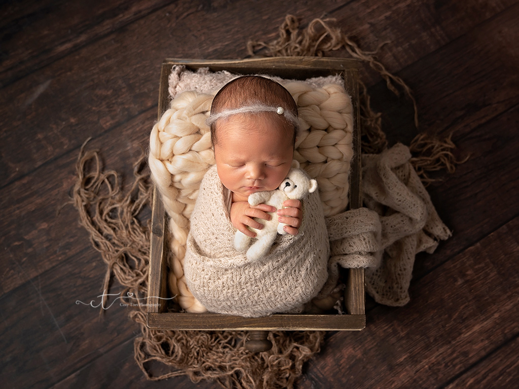 newborn baby girl swaddled holding teddy asleep in wooden crate at newborn photoshoot East Grinstead