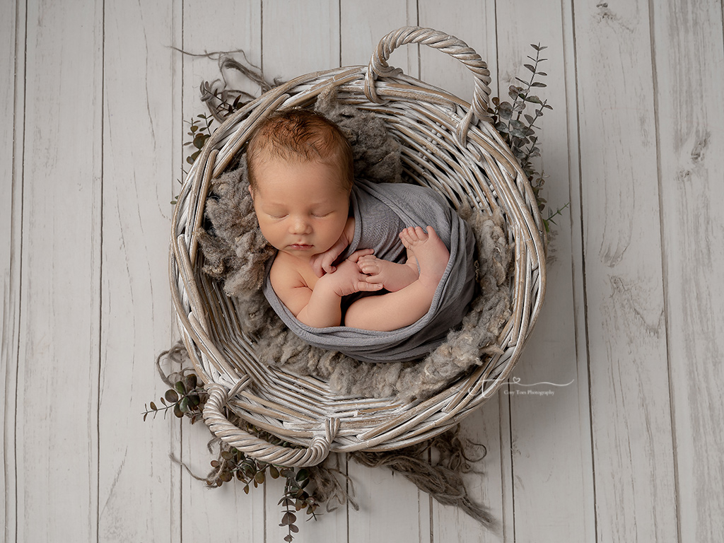 newborn baby boy swaddled and sleeping in wicker basket at his newborn photoshoot crawley