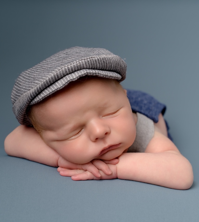 Baby in hat lying down with head resting on hands by Forest Row photography studio