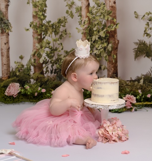 Baby wearing small crown eating cake surrounded by flowers by East Grinstead photographer