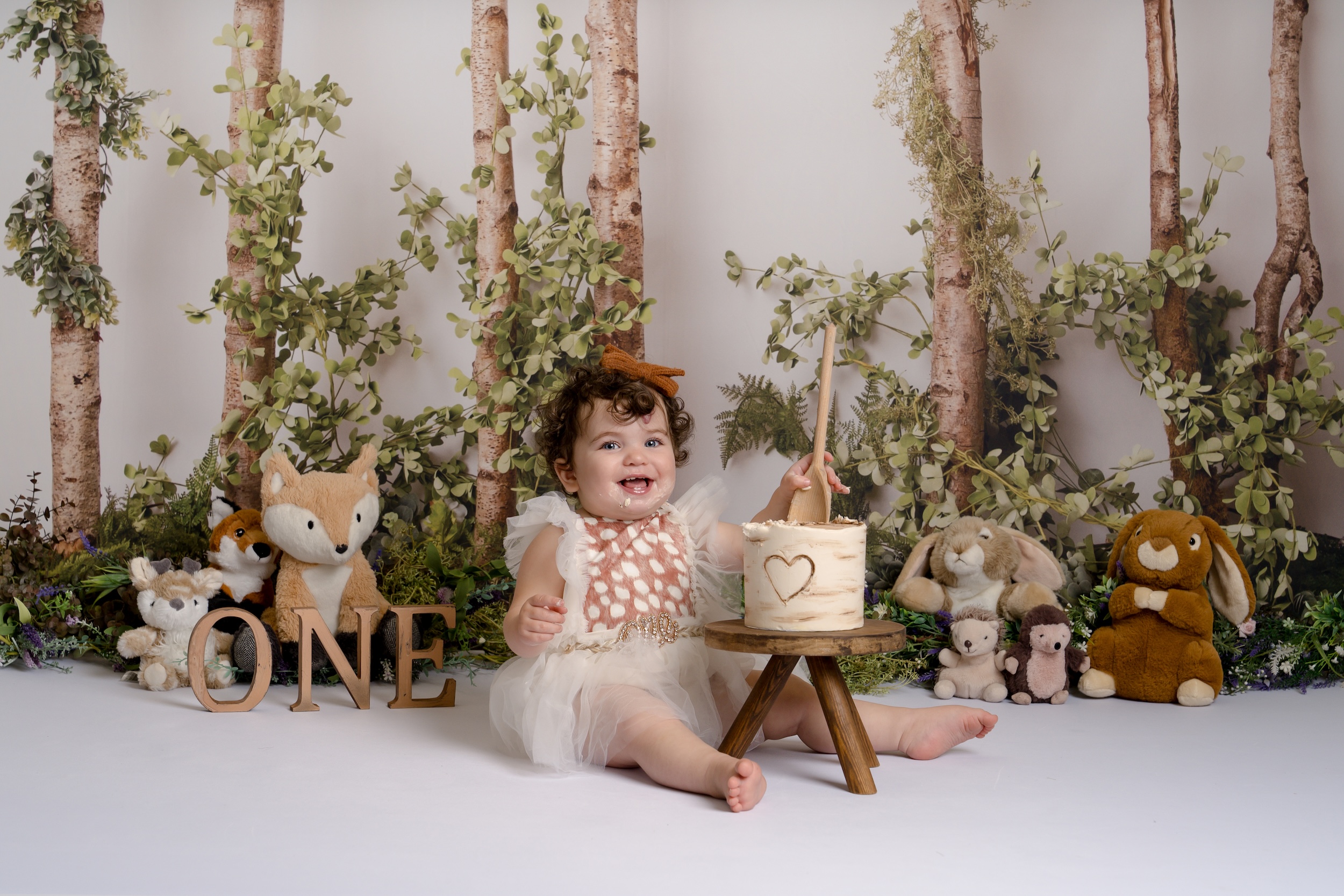 Baby holding a wooden spoon in a cake in front of stuffed animals and a tree background by Crawley photography studio