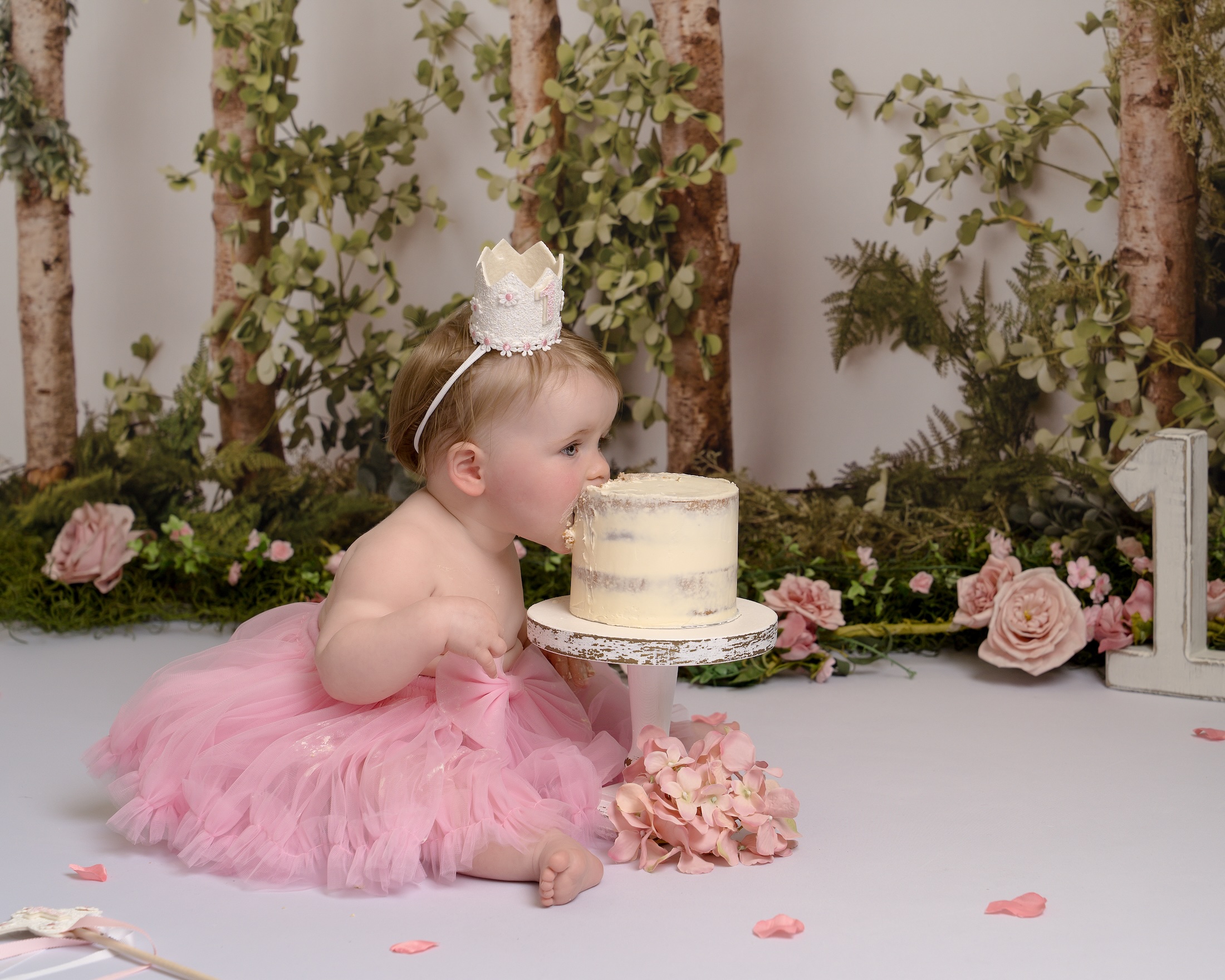 Baby wearing small crown eating cake surrounded by flowers by East Grinstead photographer