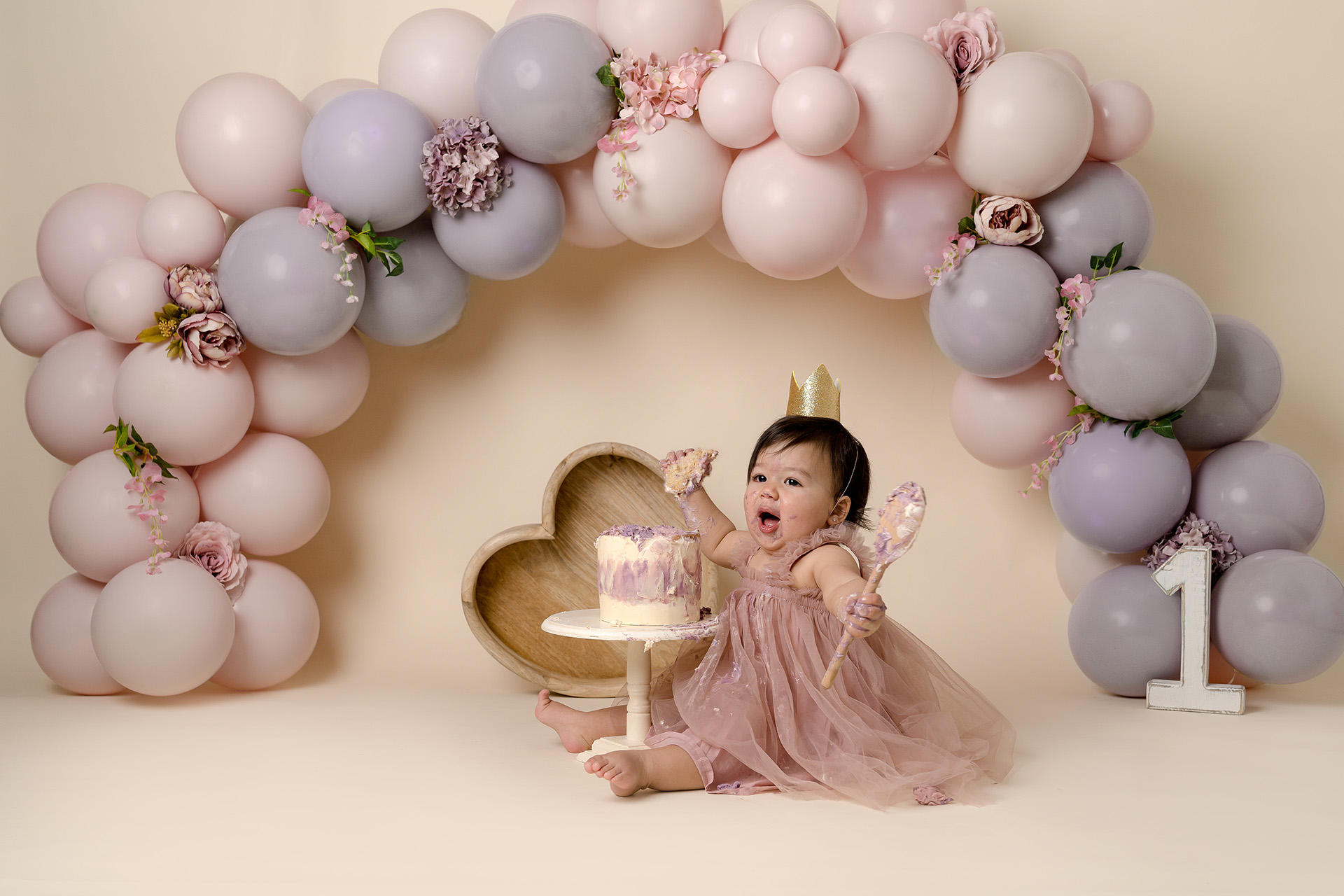 Baby girl looks excited holding wooden spoon with a baloon garland in the background by photography studio neat Redhill