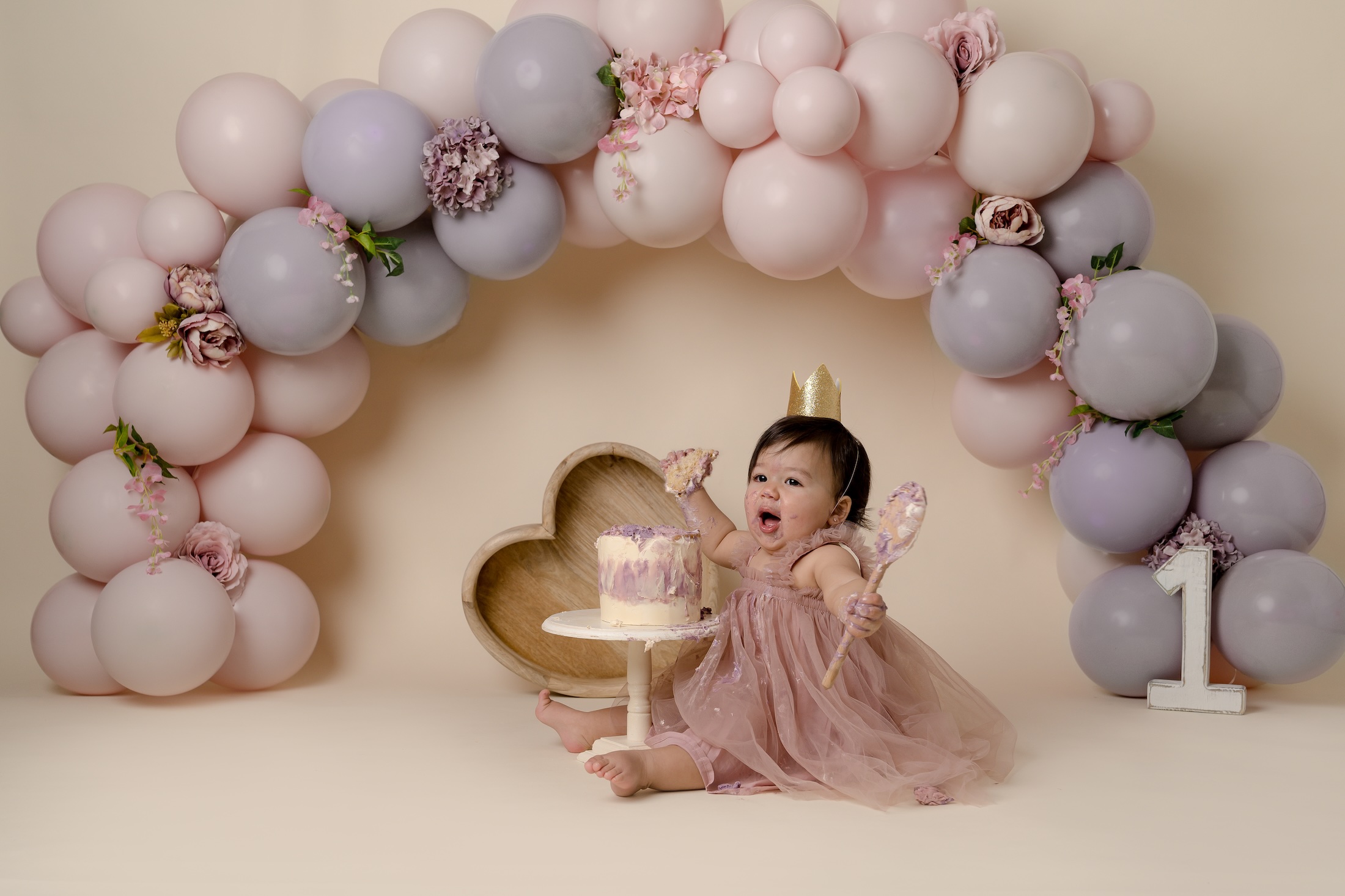 Baby girl looks excited holding wooden spoon with a baloon garland in the background by photography studio neat Redhill