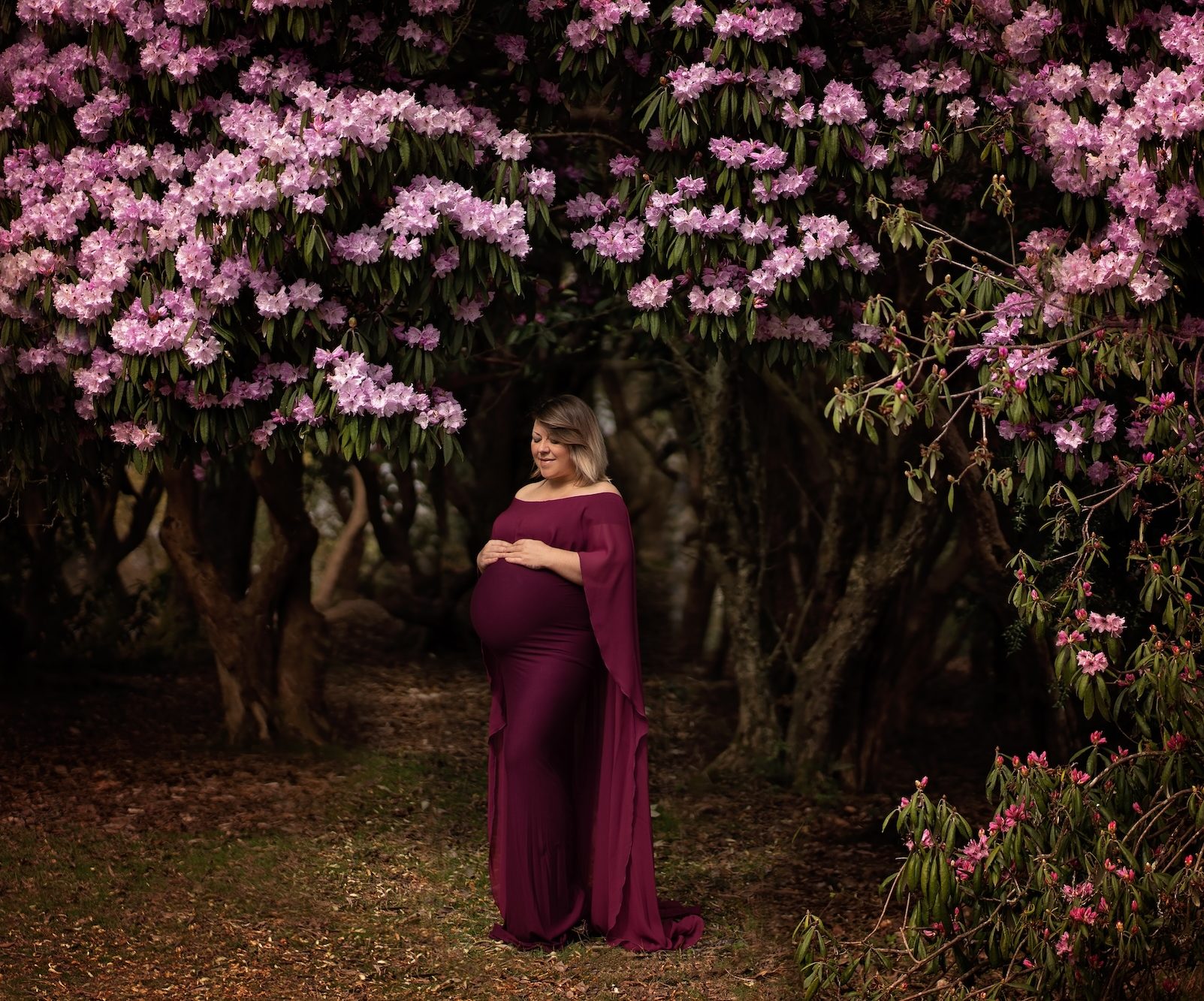 Lady holding belly under pink flowers in maternity shoot by Horsham photographer