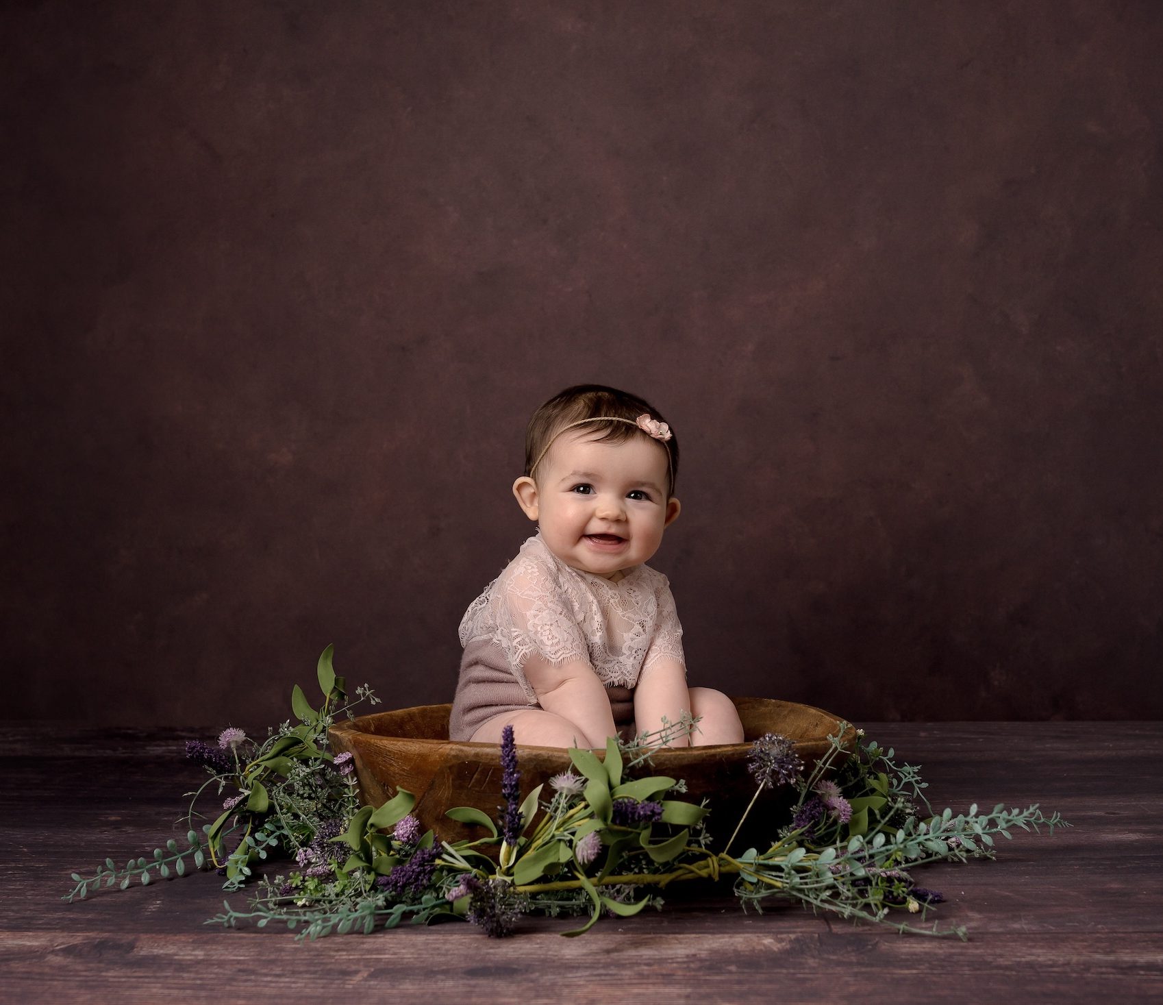 Baby smiling with flower headband sitting in wooden bowl surrounded by plants
