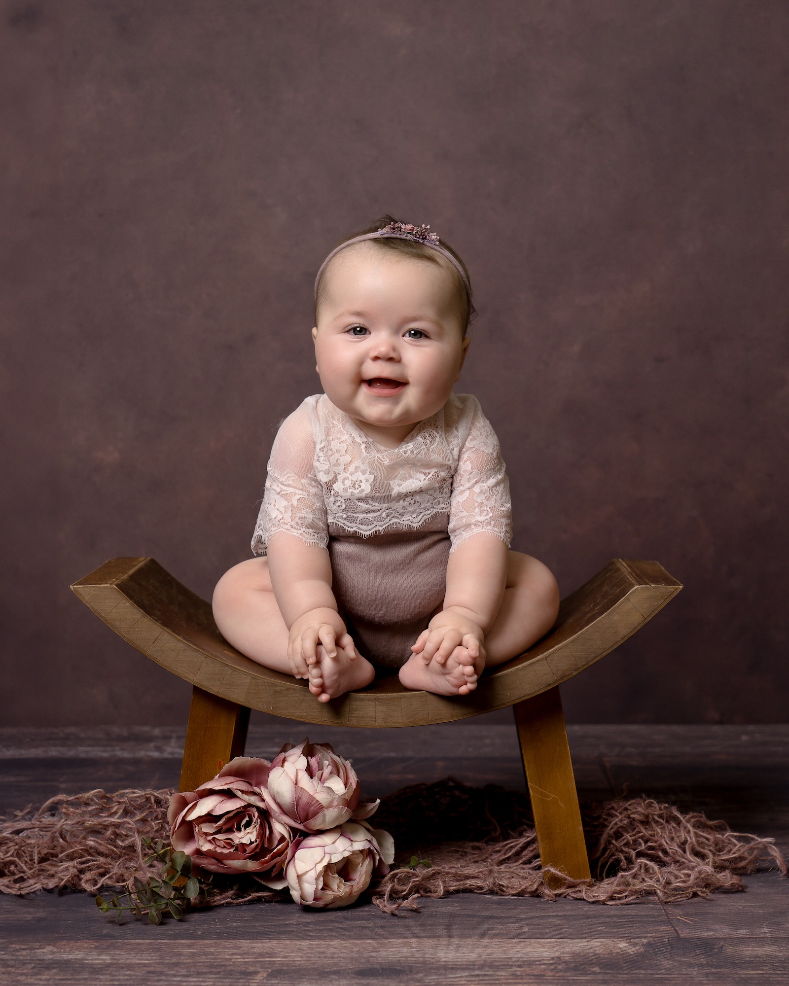 Baby sitting on wooden stool with decorative flowers and fabric on floor below by Sussex photography studio