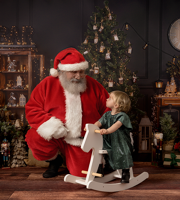 Toddler girl sitting on rocking horse looking up at Santa as she visits Santa for a photoshoot in Sussex